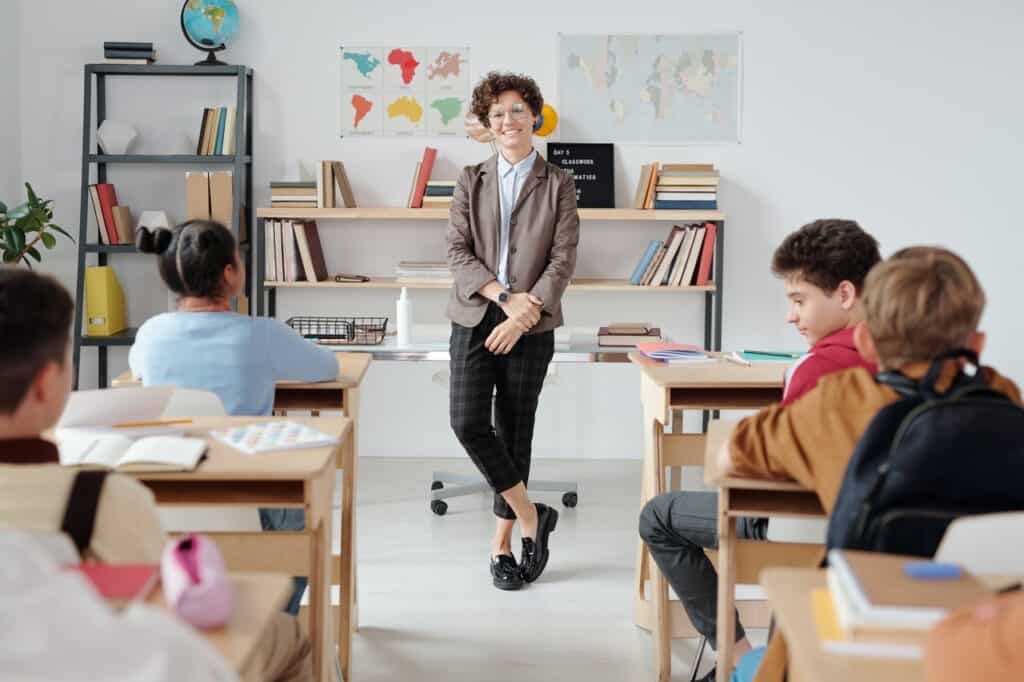 children sitting at desks looking towards the teacher leaning on her desk at the front of classroom